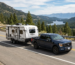 Blue pickup truck towing white GeoPro camper on mountain road with pine trees and lake